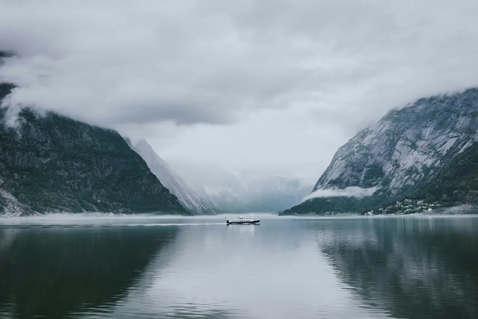 Lite passasjerskip på en stille fjord mellom tåkete fjell